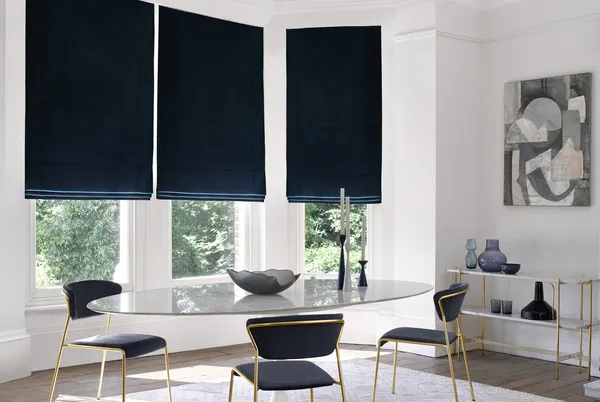 Modern dining room with bay window dressed in three navy blue velvet Roman blinds, oval glass table, metallic gold chairs with blue upholstery, metallic gold side table with blue glassware.