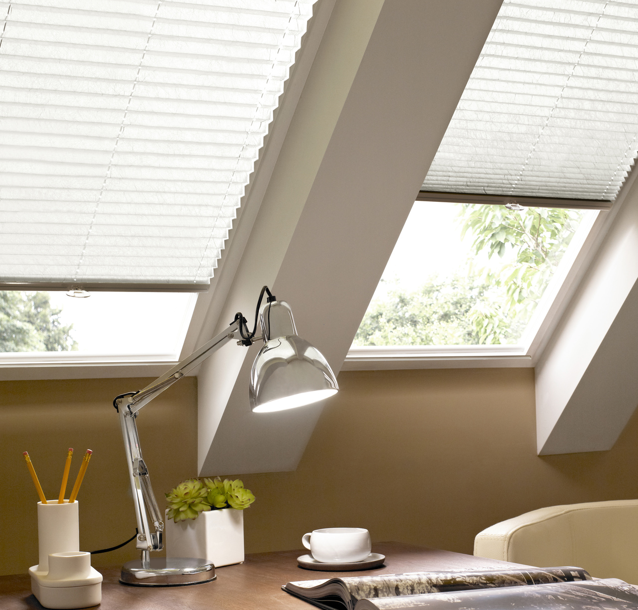 Close up on eaves with desk beneath two skylight windows dressed in off-white pleated blinds one lowered halfway, the other three quarters, silver anglepoise lamp and small plant in white pot, white desk tidy with pencils and a white cup and saucer.