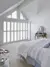 Bedroom with white bedroom gable end windows and doors with closed white shutters, louvres open, alongside a wooden vanity table and in foreground part of a bed dressed in white and pale blue bedding.