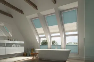 Large bathroom with sloped ceiling with three skylight windows and three standard windows directly underneath, top three dressed in twinshade duette blinds, one pale blue one cream, blue lowered to halfway cream almost to bottom, lower windows blue blinds cafe style, a white bathr, walk-in shower and white sink units.