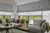 Large lounge with windows and doors on two sides with grey carpet and corner sofa in grey windows dressed in white sheer roller blind with grey patterned roller blind on top.
