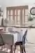 White kitchen with window behind sink dressed in pale pink-salmon shutters closed, with louvres open, cream units, wooden table with pale blue tablecloth and runner with white china.
