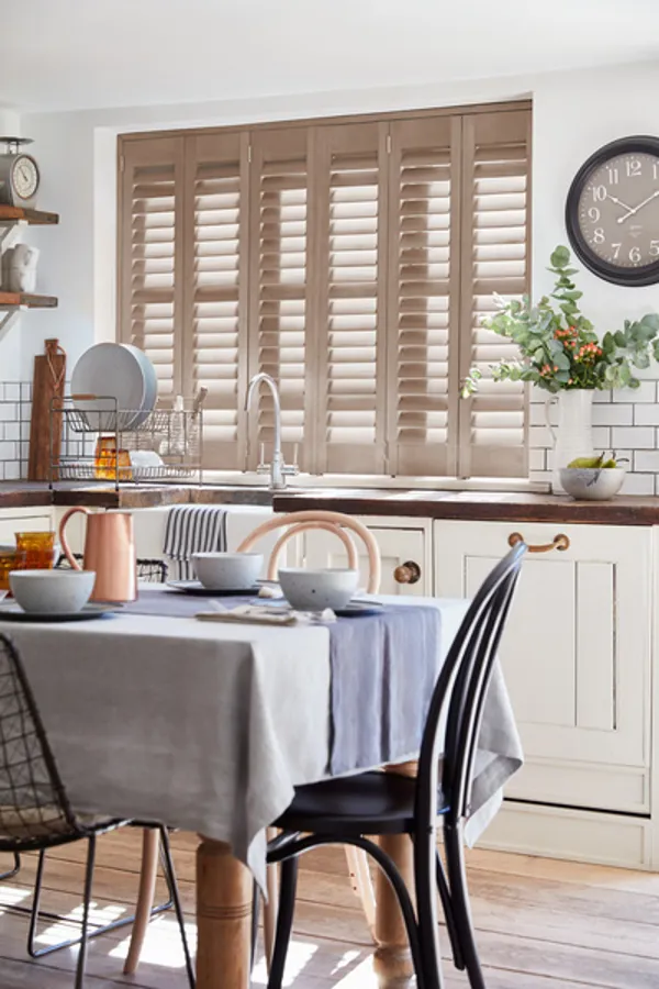 White kitchen with window behind sink dressed in pale pink-salmon shutters closed, with louvres open, cream units, wooden table with pale blue tablecloth and runner with white china.