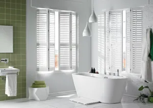 White bathroom with one walled tiled in olive green with window either side of corner dressed in white shutters, partially open above a bath.