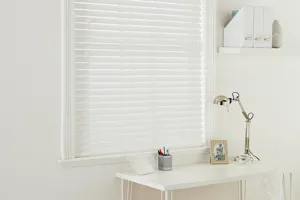 White room with a white work desk, metallic anglepoise lamp in front of a window dressed with a white faux wood venetian blind, fully lowered.