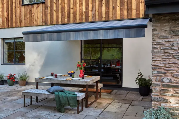 Exterior of split render and wood panel housewith grey striped awning extended over wooden table and benches set for meal, cushions and throws on bench.