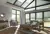 Grey living room with wood floor, grey sofa and chair, wood table and coffee table, gable end windows and doors, glass roof and patio door to left dressed in pale pine green duette blinds at varying levels.