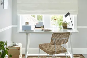 Pale grey room with modern white work desk with stationery items, raffia and metal chair all in front of a window dressed with an ivory Roman blind, lowered half way.