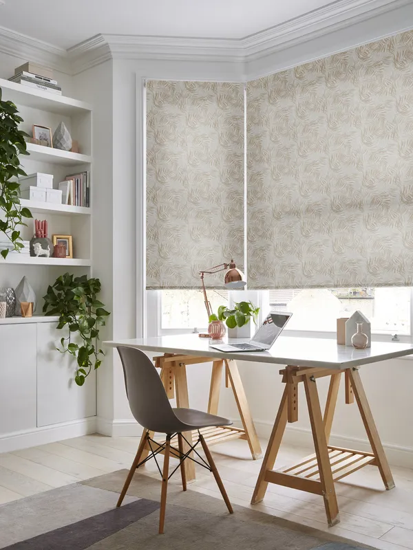 Home office with pale wood floor and large abstract pattern rug, trestle table desk with small angle poise lamp over laptop in front of bay windows dressed in roller blinds with a pale brown swirling pattern.