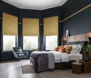 Large bedroom with wooden floor, bay window with three ochre faux velvet Roman blinds, navy walls, bed with white bedding and grey quilted throw,. 