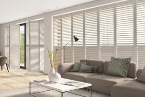 Large lounge diner with two sets of multiple door-window combinations dressed in white tracked shutters, one set partially open. Contemporary couch to one end, contemporary dining table and chairs at other.