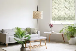 Lounge in neutral colours with cream rug, mink sofa to left, cream lamps, two large planters with ferns and window dressed in Roman blind in pale green with green fern pattern.