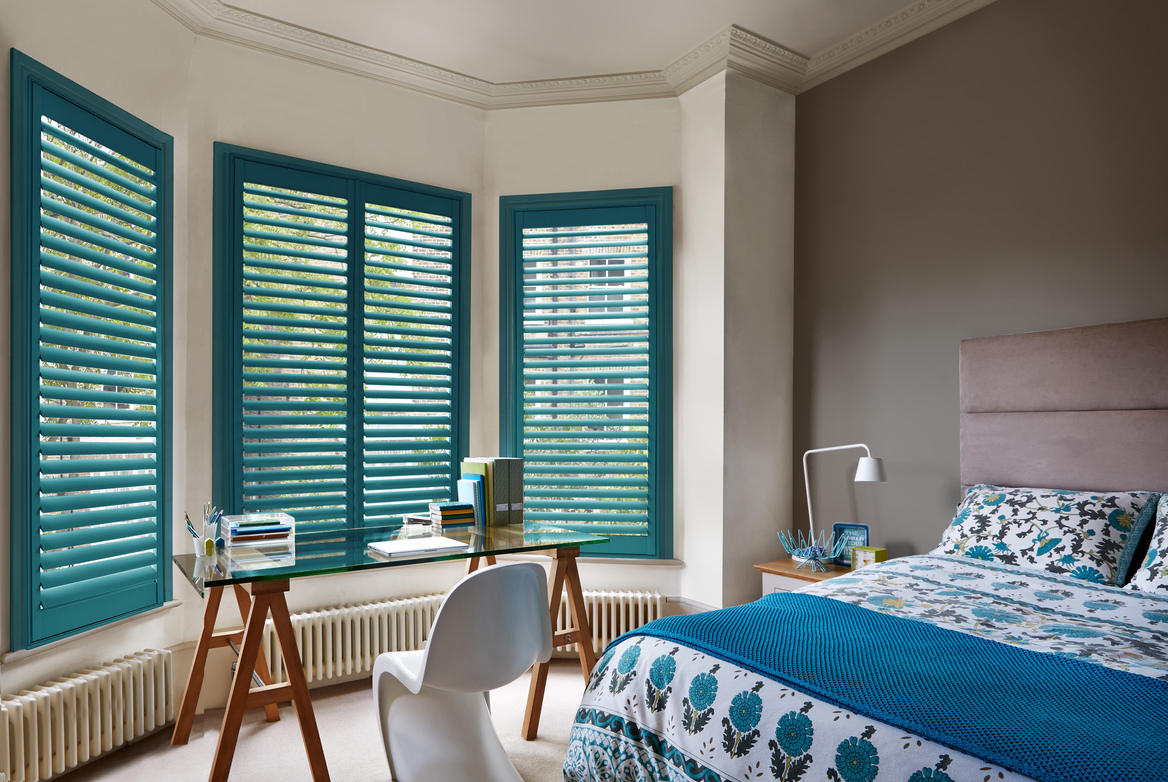 Bedroom with  bay window dressed in teal shutters, louvres open, mink wall to right behind bed with mink headboard with white and teal patterned bedding, modern glass trestle-style table in front of window.